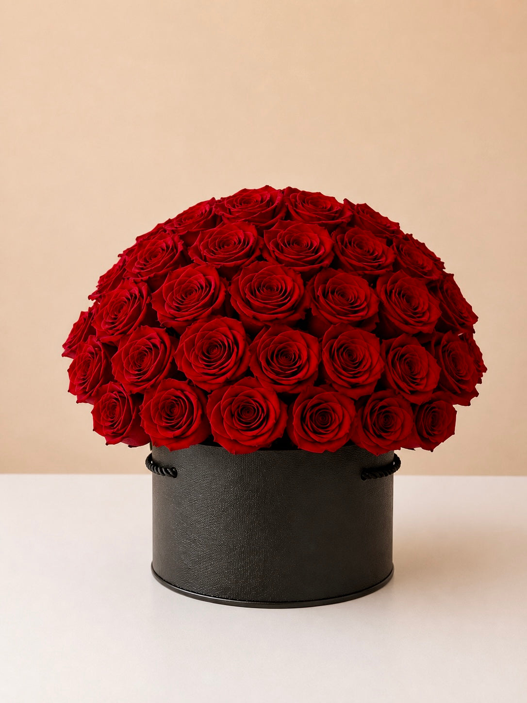 A dome-shaped arrangement of deep red roses in a round black box, photographed on a clean white surface against a warm beige background.